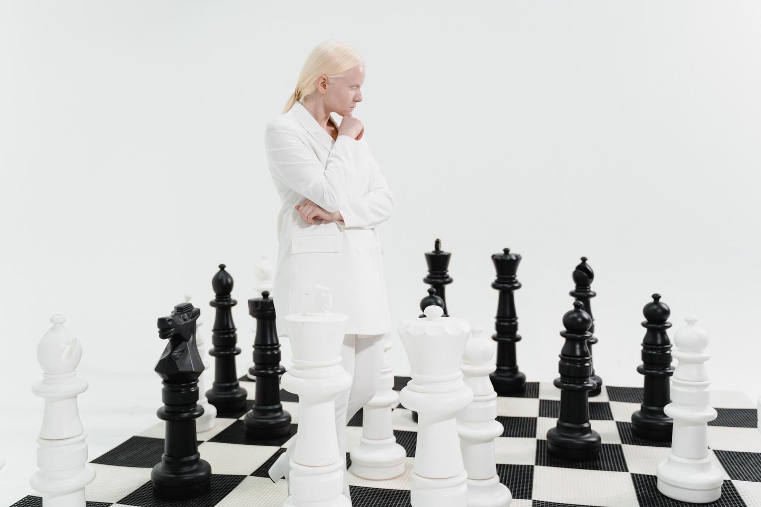 Mujer contemplando piezas de ajedrez gigantes en un tablero, simbolizando la estructura y las reglas del pensamiento ajedrecístico. Mujer contemplando piezas de ajedrez gigantes en un tablero, simbolizando la estructura y las reglas del pensamiento ajedrecístico.